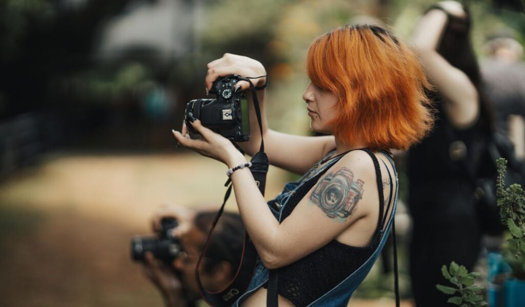  Woman with bright orange hair and a camera tattoo on her arm taking a photo with a DSLR camera outdoors.