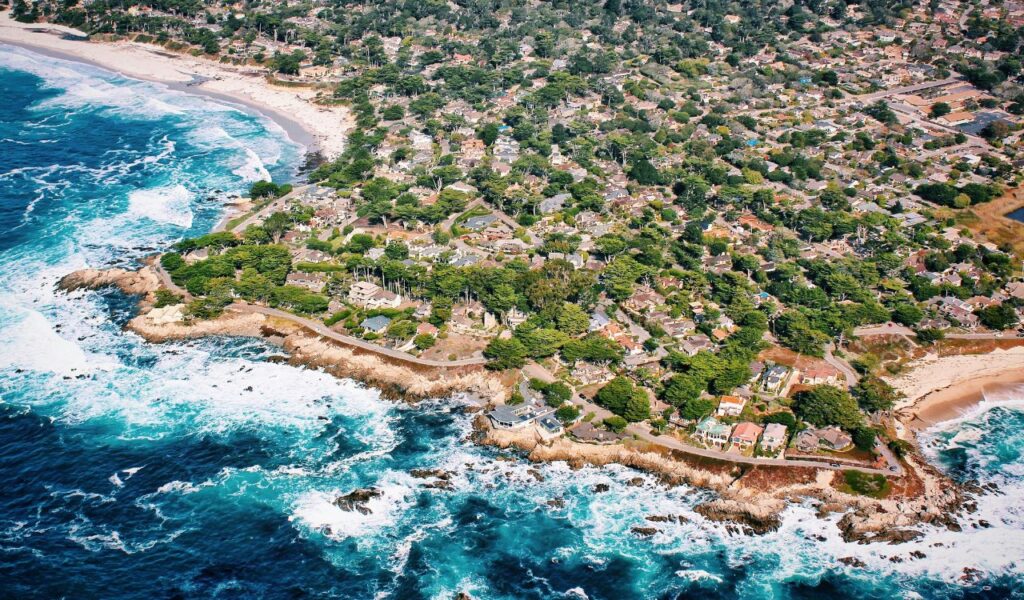 Aerial view of Carmel-by-the-Sea, California, showing a coastal neighborhood with houses nestled among trees, rugged cliffs, and waves crashing along the Pacific shoreline.