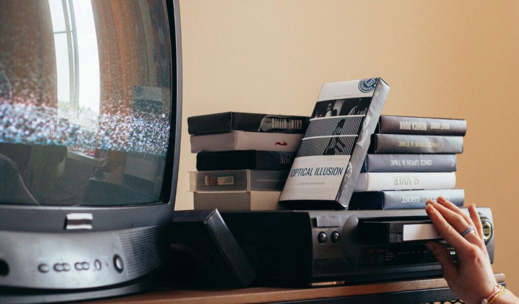 A person inserts a VHS tape into a player next to a CRT television, with stacks of tapes and a screen showing static in the background.