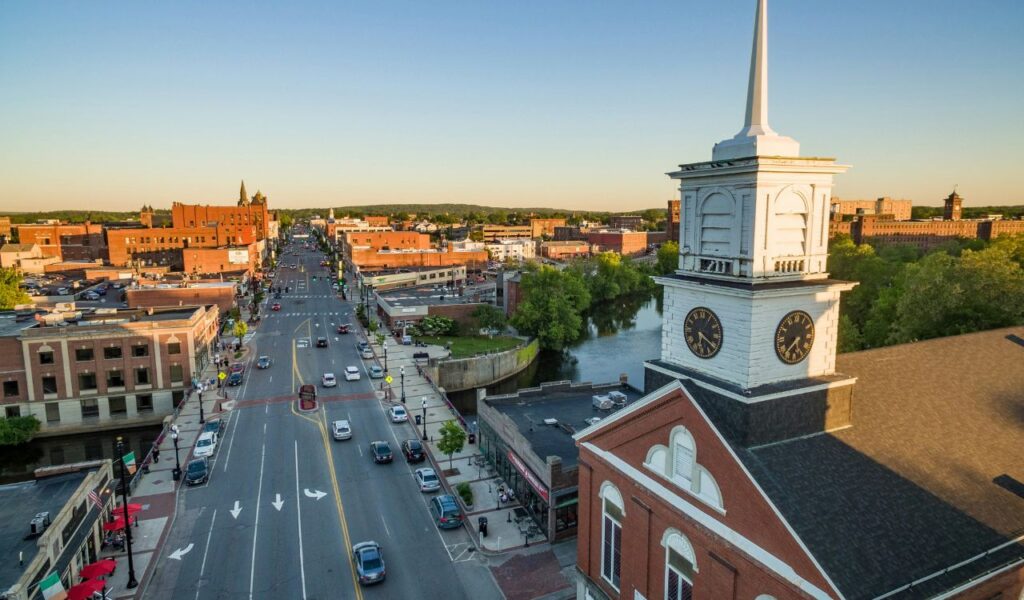 Aerial view of Central Square in Keene, New Hampshire, featuring a prominent white clock tower, historic brick buildings, a main street lined with cars, and a river curving through the town under a clear evening sky.