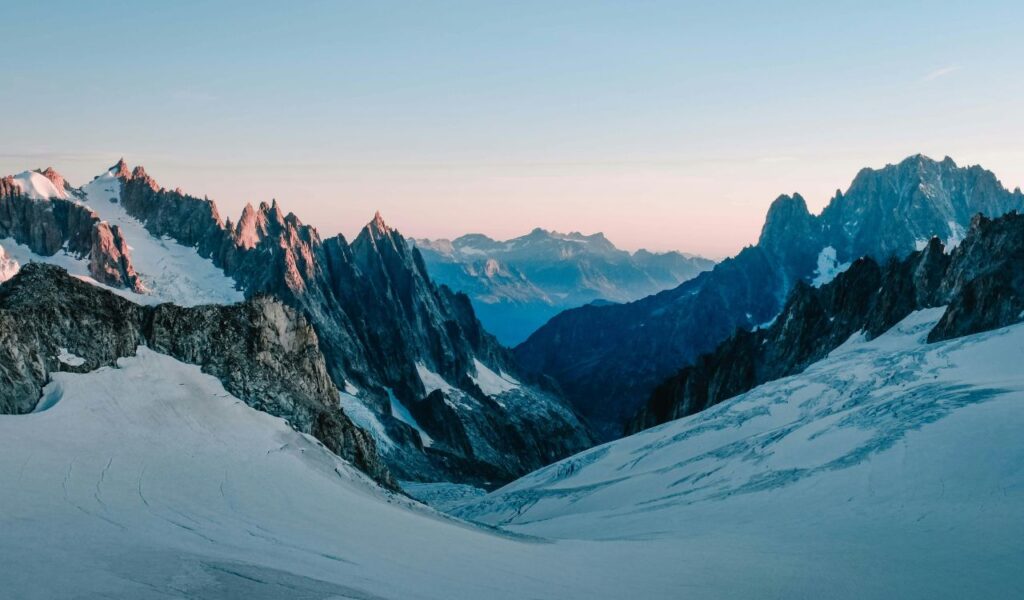 Breathtaking view of snow-covered peaks and sharp ridges in Chamonix, France, with glowing light from the setting sun casting a soft pink hue over the rugged alpine landscape.