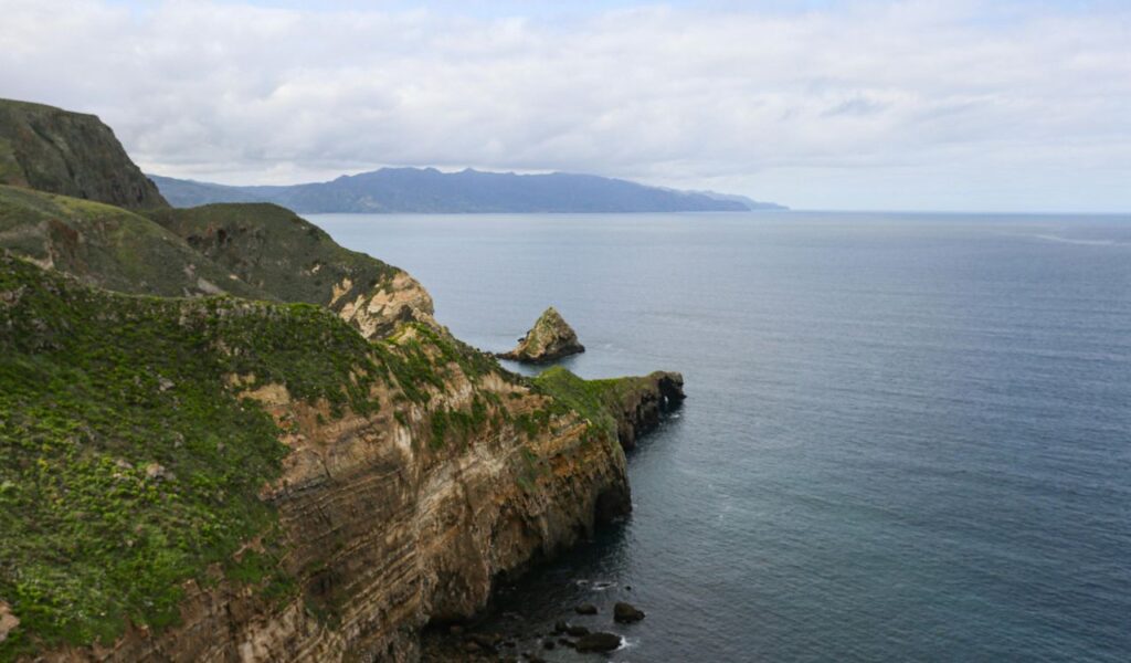 A scenic view of rugged green cliffs extending into the calm blue ocean at Channel Islands National Park, with distant mountains visible under a partly cloudy sky.