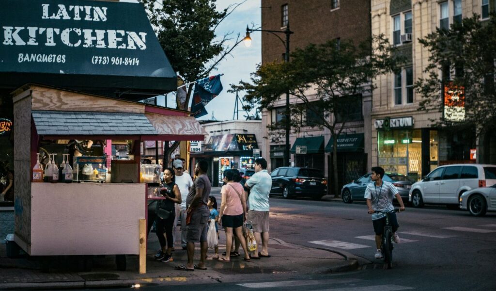  Evening street scene in Chicago with people gathered at a small food stand under a sign reading "Latin Kitchen," surrounded by storefronts, parked cars, and a cyclist passing by.