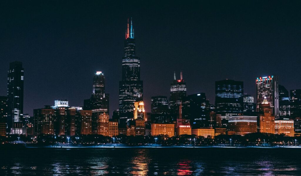 Chicago skyline at night with illuminated skyscrapers, including Willis Tower, reflecting vibrant colors onto the river.
