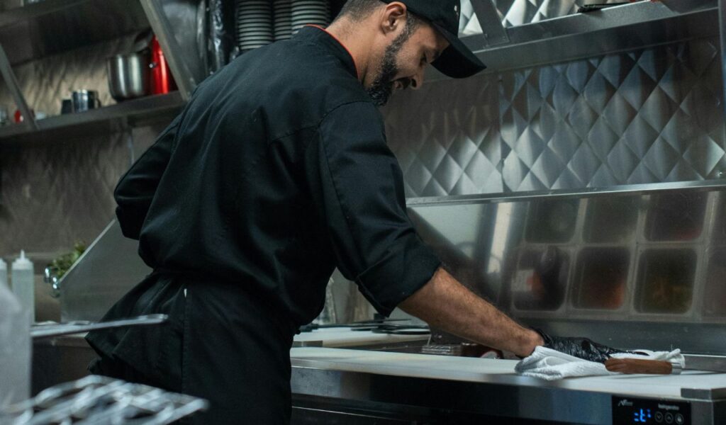 A chef in a black uniform cleans a stainless steel kitchen counter, ensuring equipment and surfaces are properly sanitized.