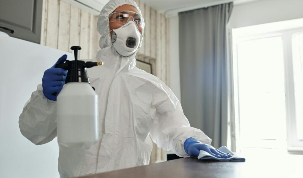A person in protective gear and gloves sanitizes a surface with a spray bottle and cloth, ensuring thorough cleaning and hygiene.