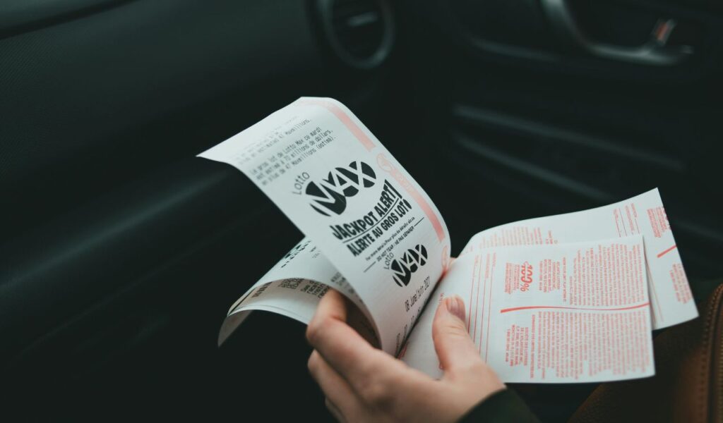 A person is holding a Lotto Max ticket booklet inside a car, with the "Jackpot Alert" text visible on the ticket, focusing on lottery details.