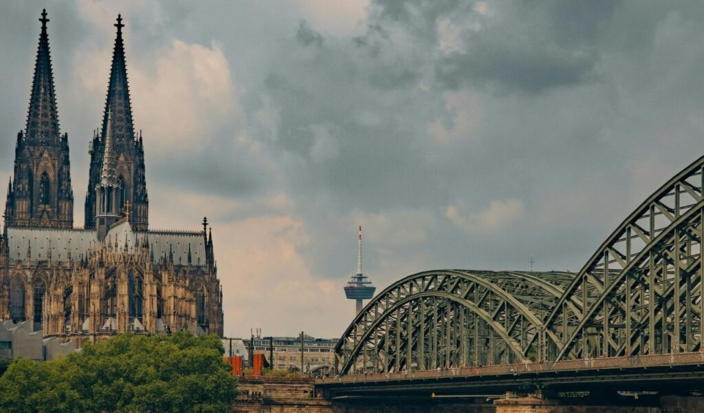 Cologne Cathedral in Germany, with its two towering Gothic spires beside the Hohenzollern Bridge, under a cloudy sky with the cityscape and telecommunications tower in the background.