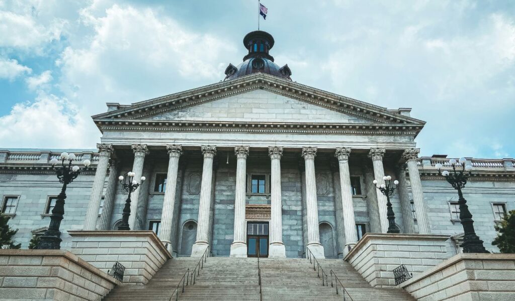 Front view of the South Carolina State House in Columbia, featuring grand stone steps, tall columns, and a dome topped with a flag.