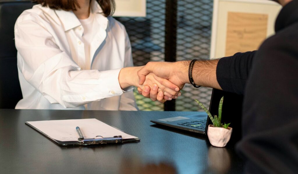 Two people shaking hands across a desk, with a clipboard, pen, laptop, and small potted plant in view, suggesting a formal agreement or financial arrangement.