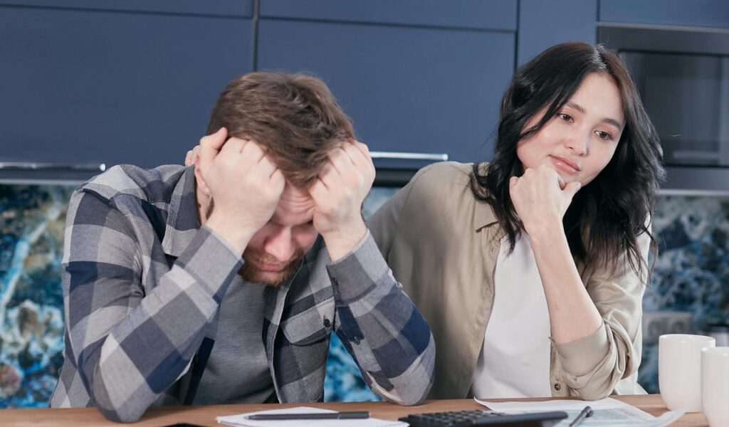 A man holding his head in frustration while a woman sits beside him looking thoughtful, with papers and mugs on the table
