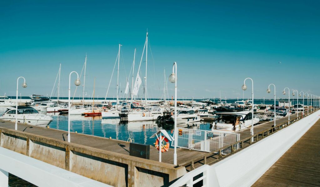 A marina filled with docked yachts and sailboats, with a wide wooden pier lined with white lamp posts extending alongside the harbor on a clear, sunny day.