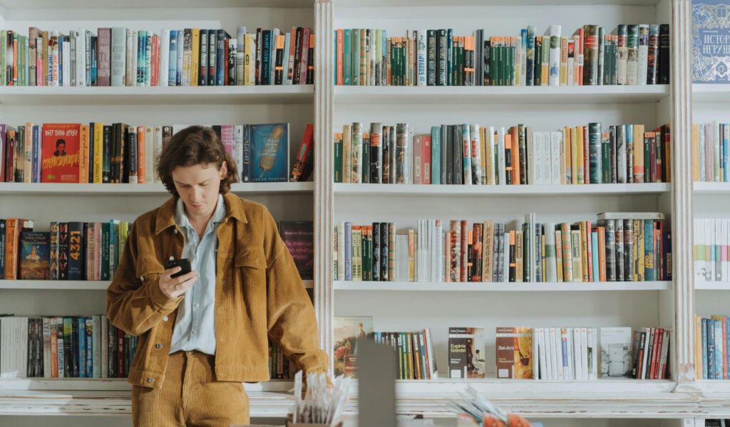A man in a brown jacket stands reading his phone in front of white bookshelves filled with colorful books at Cook & Book in Brussels.