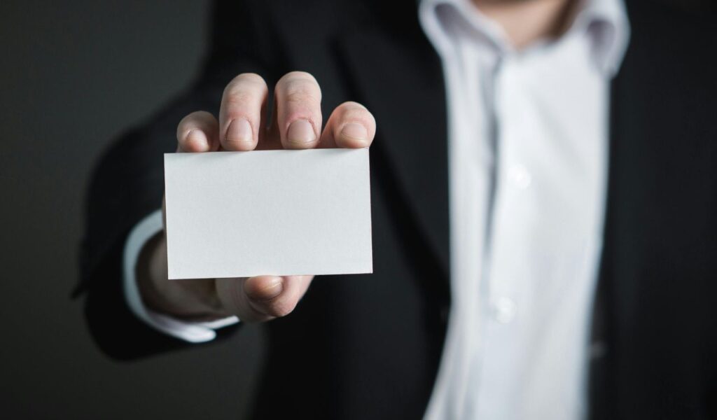 Person in a black suit holding out a blank white business card toward the camera.