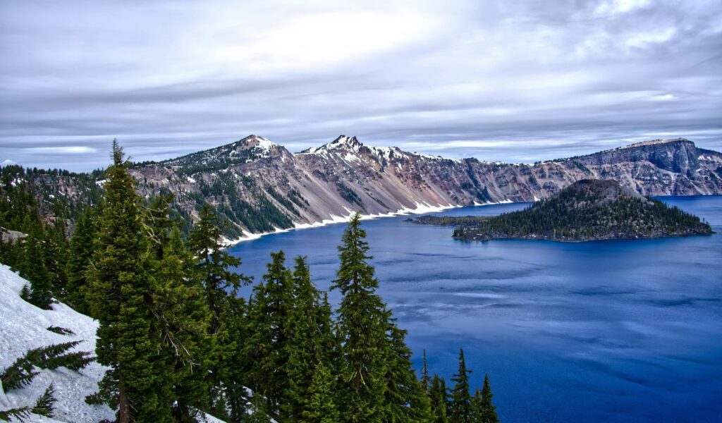 Snow-dusted cliffs and evergreen trees surround the deep blue waters of Crater Lake in Oregon, with Wizard Island in the distance.