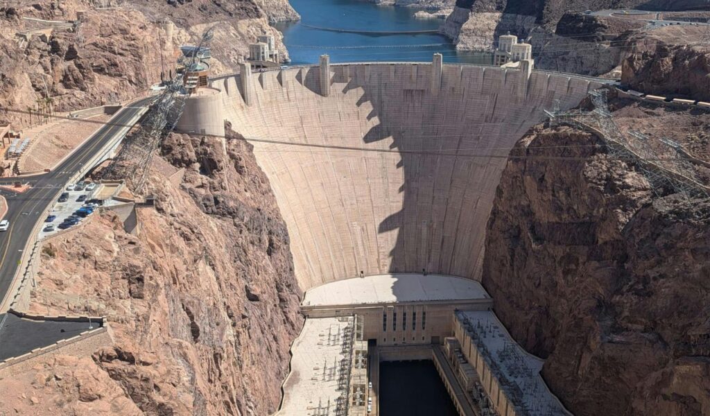 Aerial view of Hoover Dam surrounded by rocky cliffs with water visible behind the dam and clear blue sky overhead.