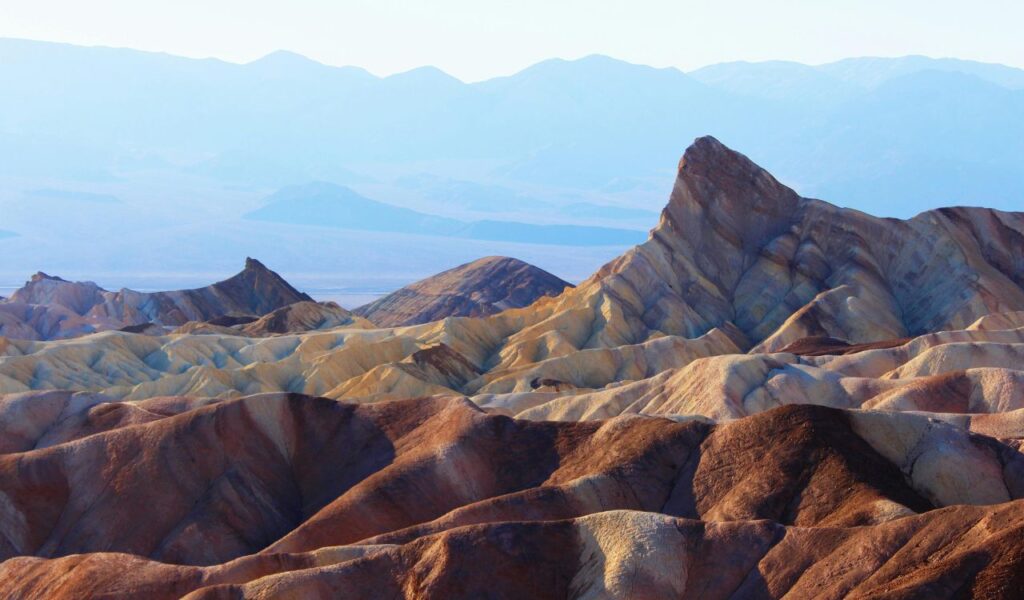 Colorful, eroded badlands and rugged peaks of Death Valley National Park with layers of mountains fading into the hazy background.