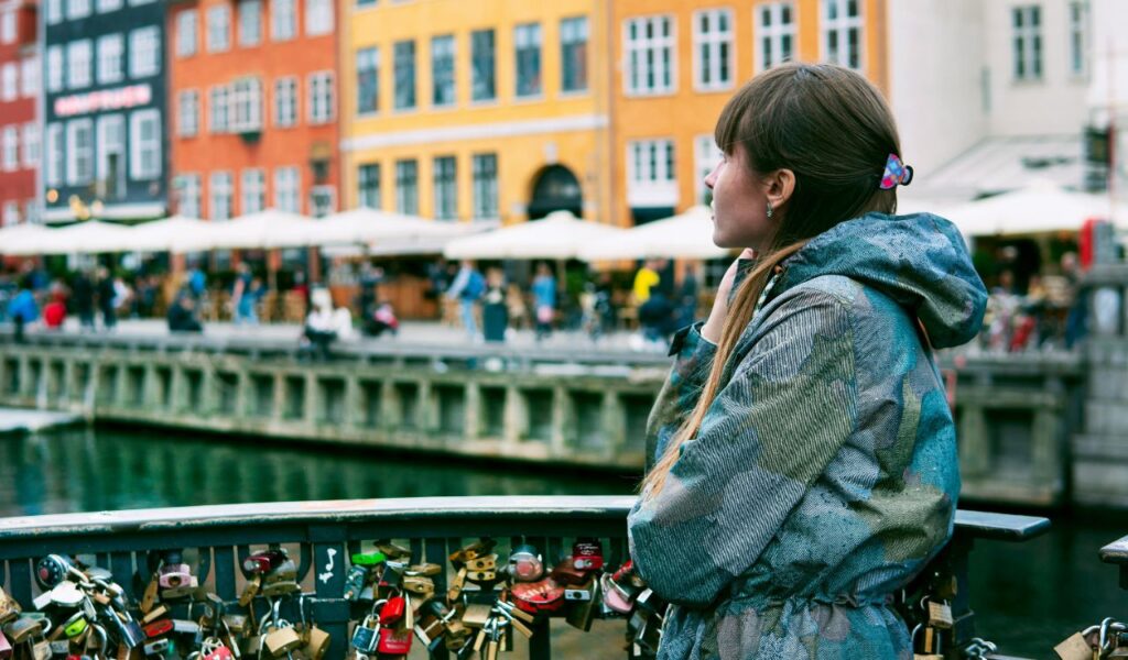 A woman in Denmark stands on a bridge decorated with love locks, gazing across the canal at Nyhavn’s colorful historic buildings and waterfront cafés.