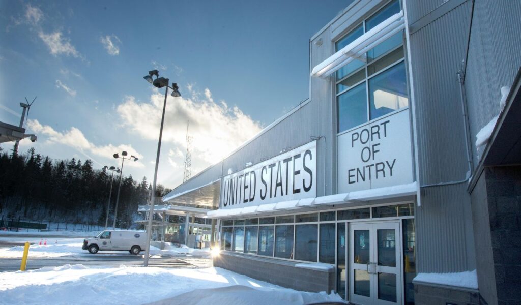 "Snow-covered United States port of entry building at a border crossing, with a van parked nearby under a clear winter sky."