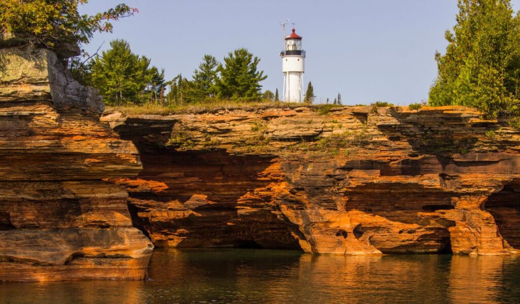 A white lighthouse rises above layered orange cliffs and still water, framed by green trees under clear skies.