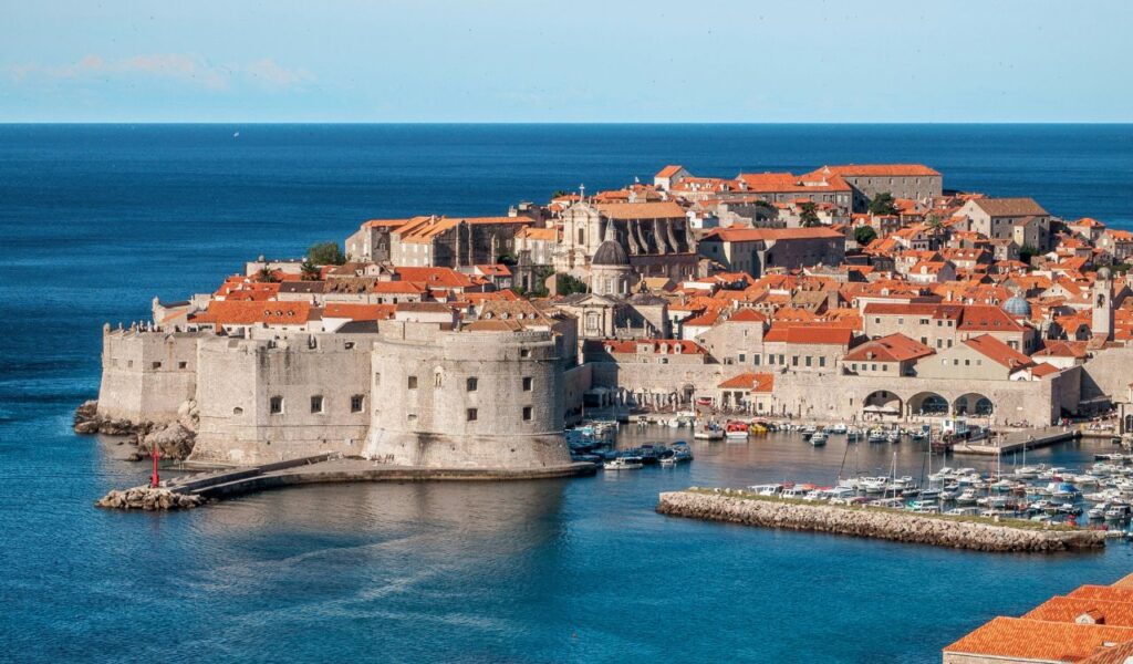 A panoramic view of Dubrovnik’s Old Town in Croatia, showcasing its medieval stone walls, red-tiled rooftops, historic buildings, and a scenic harbor against the deep blue Adriatic Sea.