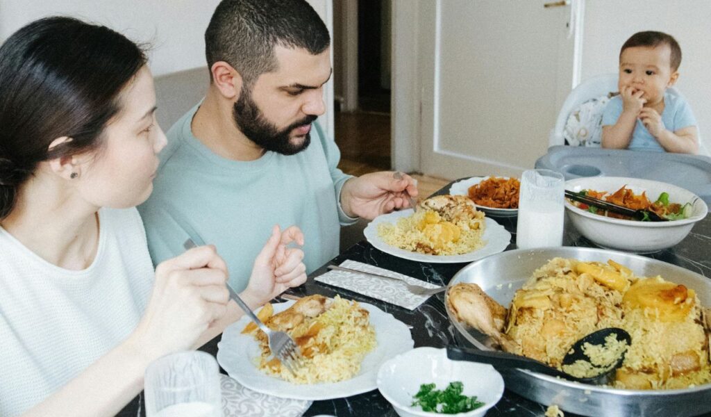 Family sitting at a dining table eating a meal of rice and chicken, with side dishes and glasses of milk.
