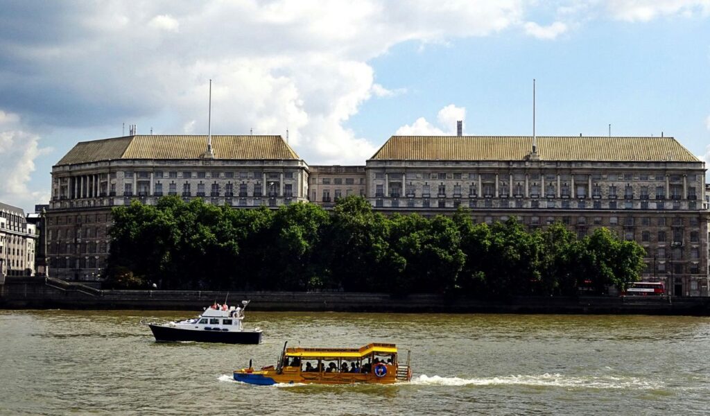 Two small boats travel along a river in front of a grand neoclassical government building lined with trees, under a partly cloudy sky