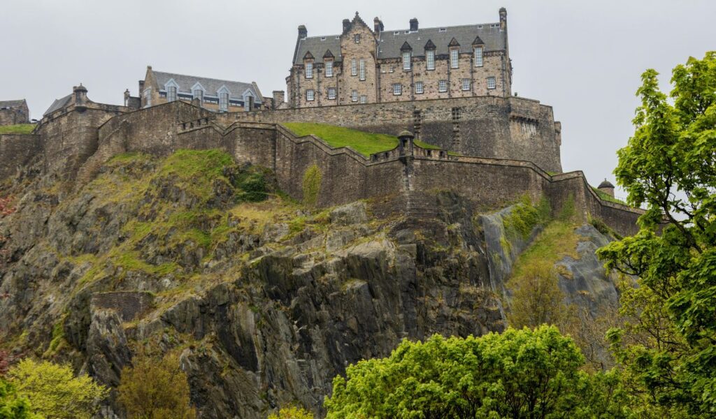 Edinburgh Castle in Scotland, perched atop Castle Rock with steep rocky cliffs, surrounded by stone walls and lush green trees.