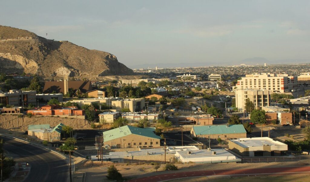 Cityscape of El Paso, Texas, with desert mountains in the background, scattered buildings, and roads under a hazy sky.
