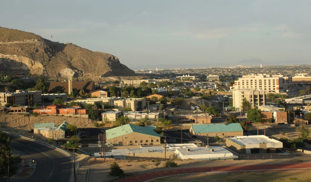 View of El Paso, Texas, with desert mountains in the background, scattered buildings, and roads under a hazy sky.