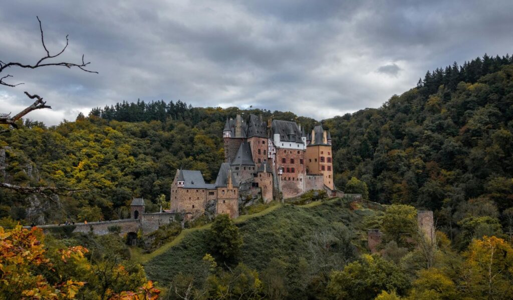 Eltz Castle in Germany, surrounded by dense green forest and autumn foliage under a cloudy sky, perched on a hill with stone walls.