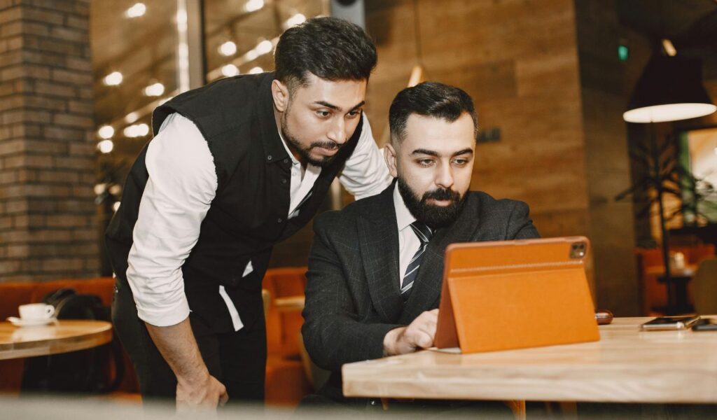 Two men in a restaurant setting, one in a vest leaning over and the other in a suit, focused on a tablet screen, possibly discussing training or safety procedures.