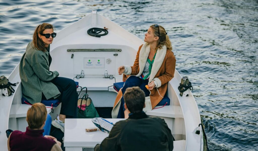 A small group of people sitting and talking on a white boat floating on calm water, with bags and personal items placed nearby.
