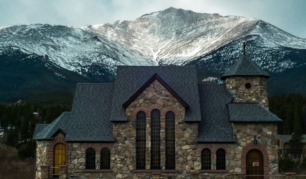 Stone church with arched windows and a turret in Estes Park, Colorado, set against snow-covered mountain peaks and a dramatic alpine backdrop.