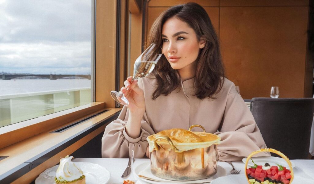 A woman sitting at a table by a large window on a cruise ship, holding a glass of white wine and enjoying a meal with elegant plating.