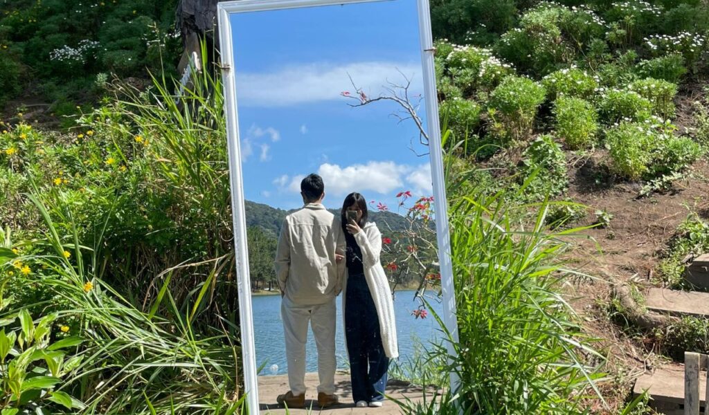  Reflection of a couple standing by a lake in a large outdoor mirror surrounded by greenery.