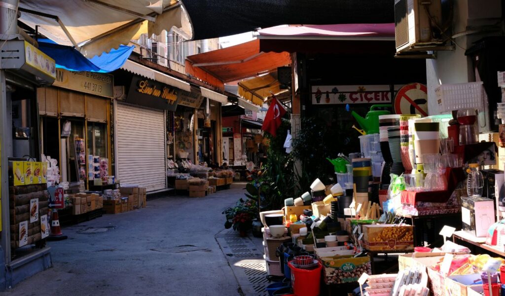 Narrow market street with shops selling household goods, plants, and packaged items, shaded by awnings and a Turkish flag hanging above.