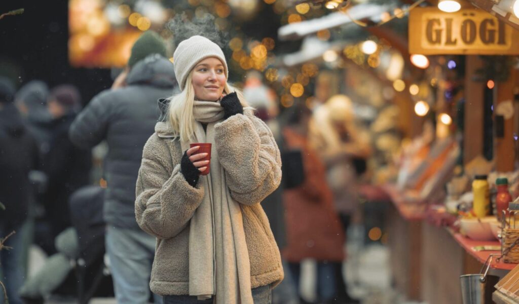 A woman in a cozy coat, scarf, and beanie enjoys a warm drink at a festive outdoor market in Finland, surrounded by glowing lights.