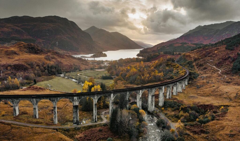 Dramatic view of the Glenfinnan Viaduct near Fort William, Scotland, curving through an autumn-colored valley with mountains and a loch in the background under a cloudy sky.