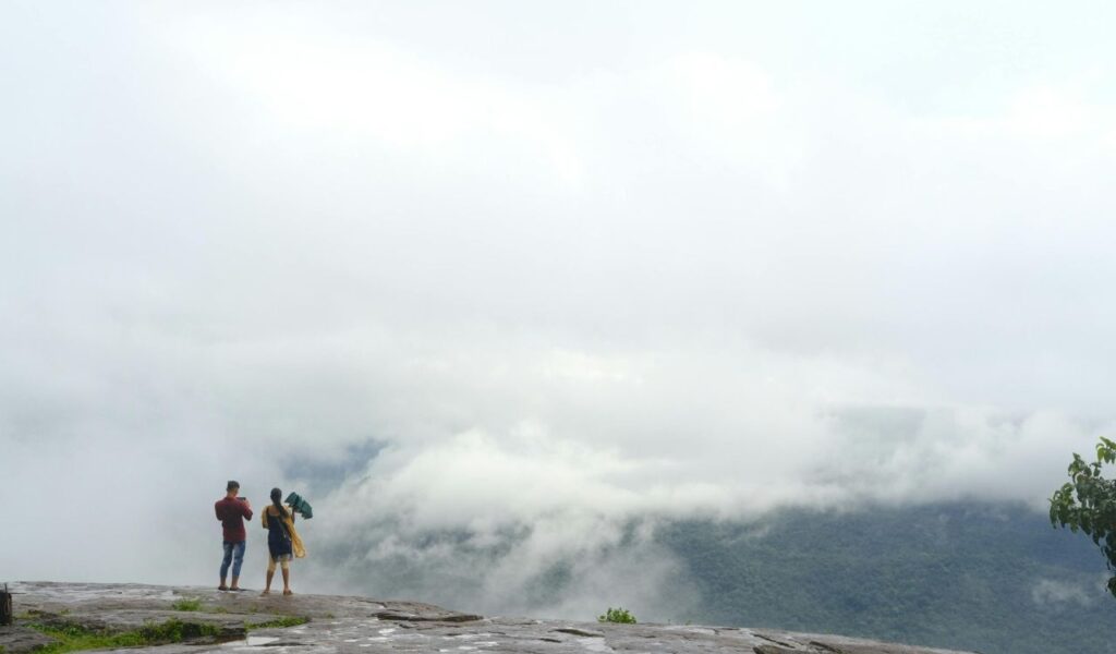 Two people standing on a rocky ledge, photographing a misty mountain landscape partially covered by clouds.