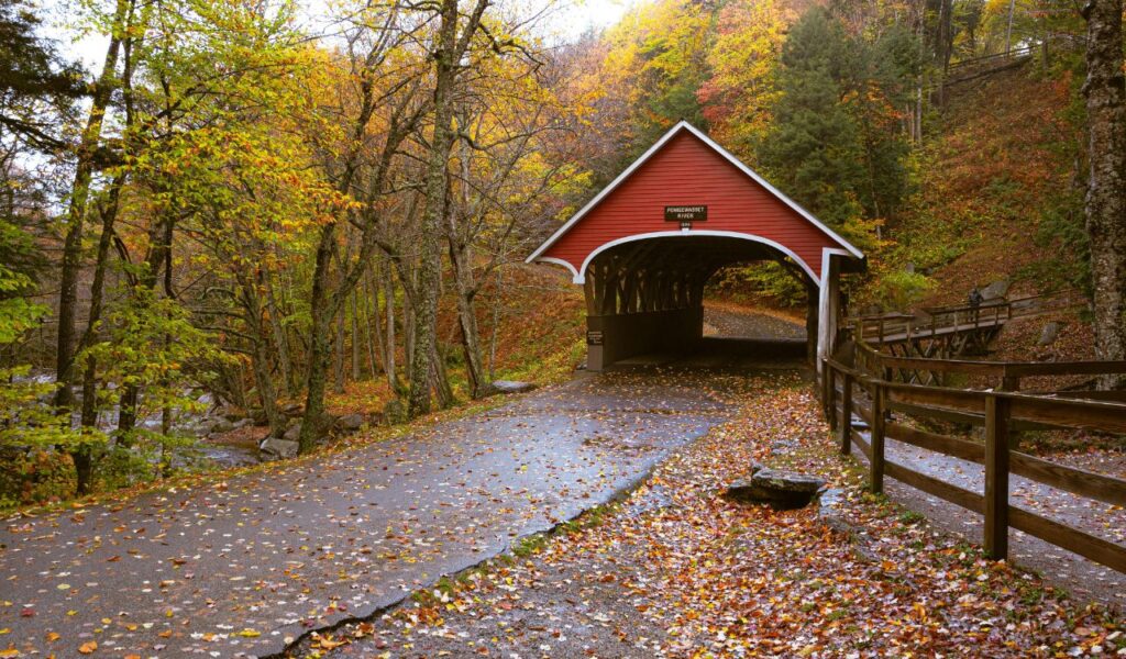 Red covered bridge labeled 'Pemigewasset River' surrounded by colorful autumn trees and fallen leaves in Franconia Notch, New Hampshire