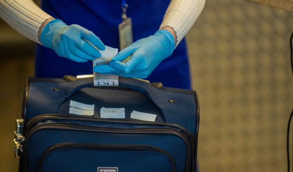 A close-up of an airport staff member wearing blue gloves attaching a luggage tag to a dark blue suitcase handle, preparing it for check-in.