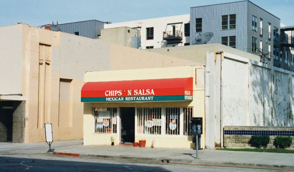 A small Mexican restaurant named Chips 'N Salsa with a red and green awning, set in a modest building along a quiet street, surrounded by larger, more modern structures in the background.