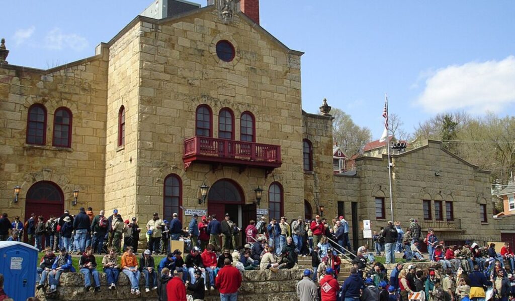 Crowd gathered outside a historic stone building in Galena, Illinois, featuring arched windows, red trim, and a small balcony, with people sitting on steps and grassy areas during a public event.