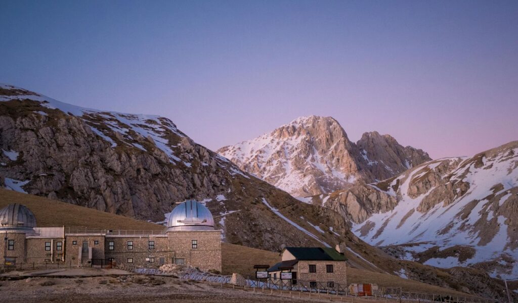 A mountain observatory with domed roofs sits at the base of rugged, snow-dusted peaks under a soft purple sky at sunset.