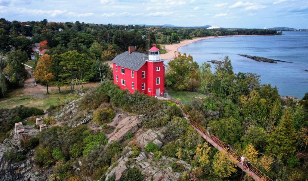 A bright red lighthouse perched on a rocky bluff surrounded by green and autumn-colored trees, overlooking a sandy beach and the calm blue waters of Lake Superior, with a narrow walkway leading to the lighthouse.