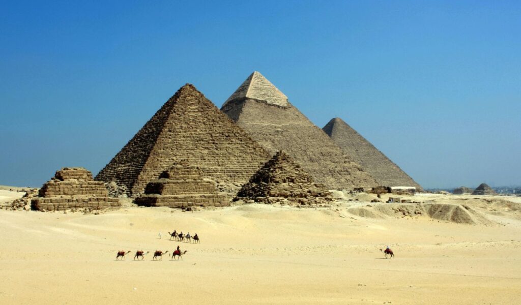 The Great Pyramids of Giza in Egypt, standing tall in the desert under a clear blue sky, with camels and riders crossing the sandy foreground.