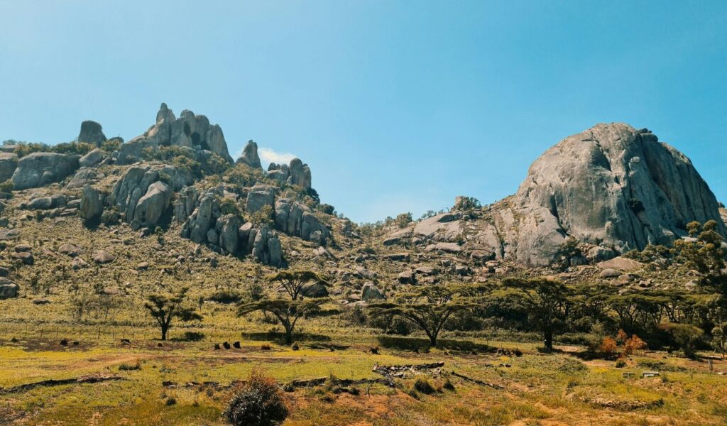 Rocky granite hills and scattered stone ruins of Great Zimbabwe, surrounded by grassy plains and acacia trees under a clear blue sky.