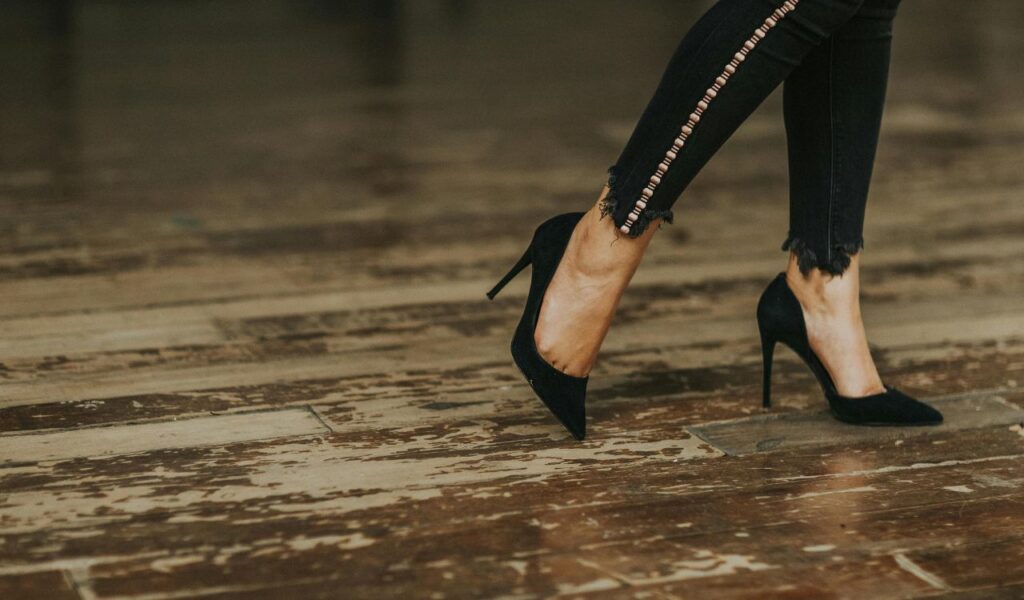 Close-up of a person wearing black high heels and black jeans walking on a wooden floor.