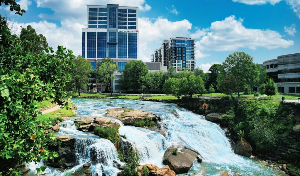 Waterfall flowing through a park in Greenville, South Carolina, with modern office buildings and trees in the background under a bright blue sky.